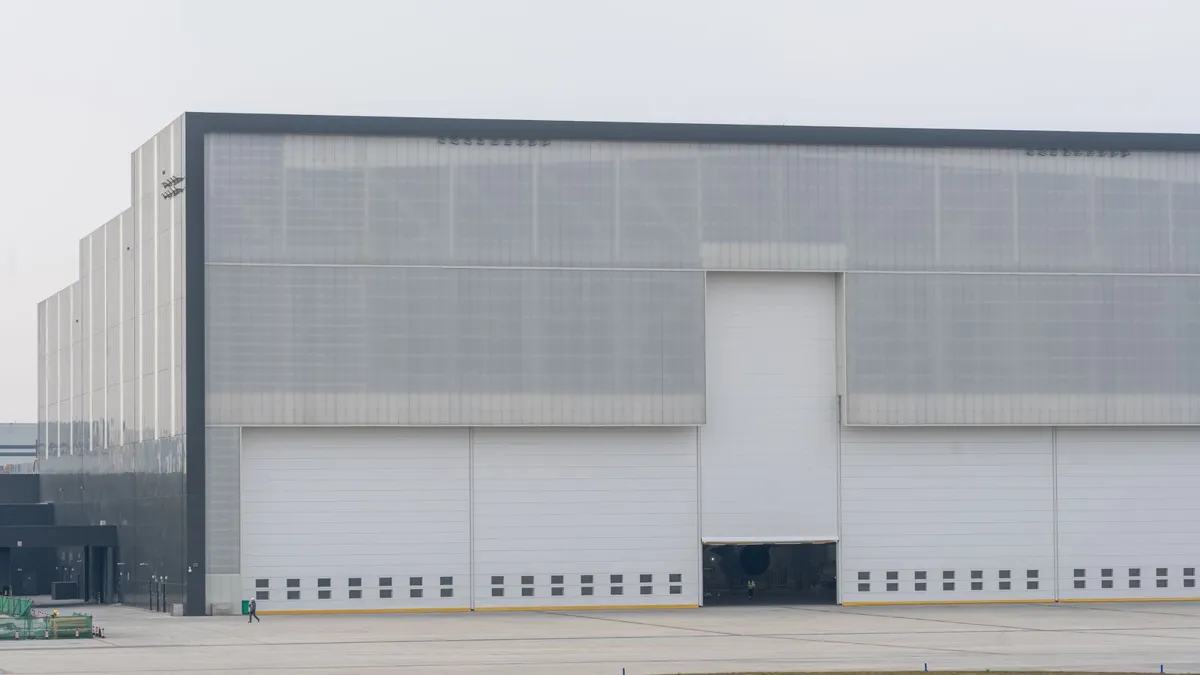 Folha de porta branca, larga e plana, inserida na fachada de um grande edifício industrial em dimensões de hangar