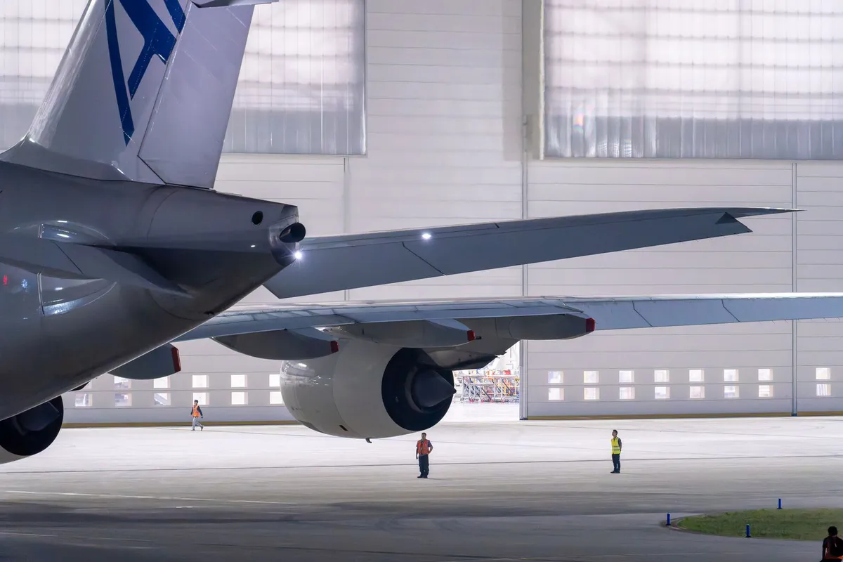 Airliner fuselage beside a wall of large paneled hangar doors with ground crew below