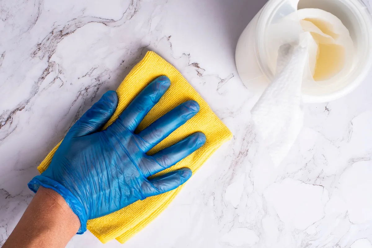 Gloved hand wiping a smooth white surface with a yellow cloth next to a disinfectant container