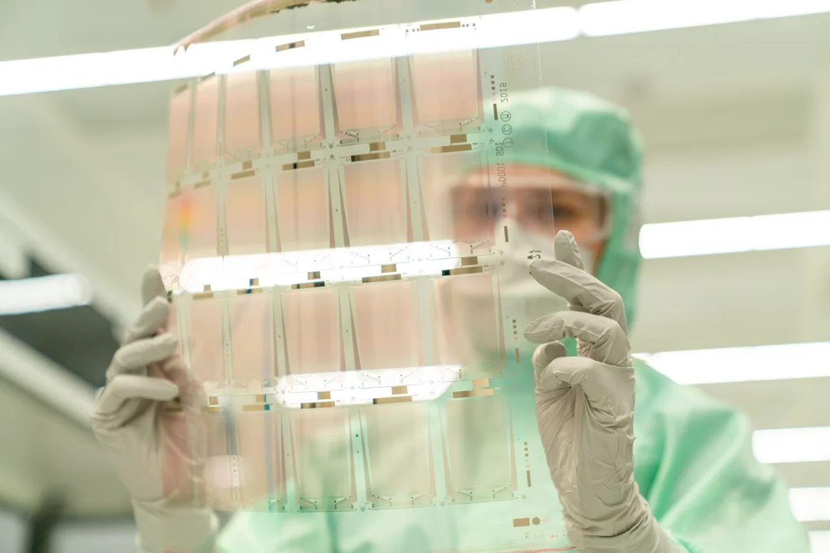 Technician in full gowning inspecting a transparent substrate panel inside a clean fabrication area